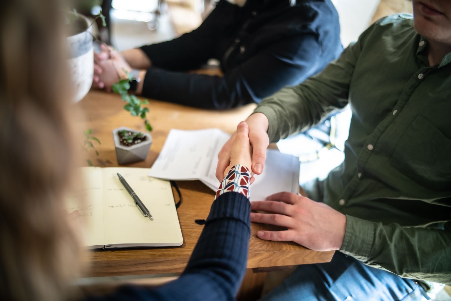 Deux personnes se serrent la main au-dessus d'une table en bois, où un carnet et des documents sont disposés dans un décor de bureau.