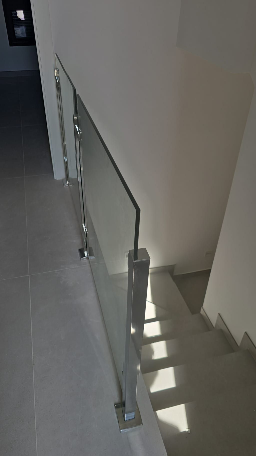Escalera con barandilla de vidrio y metal junto a un suelo de baldosas grises y una pared blanca.