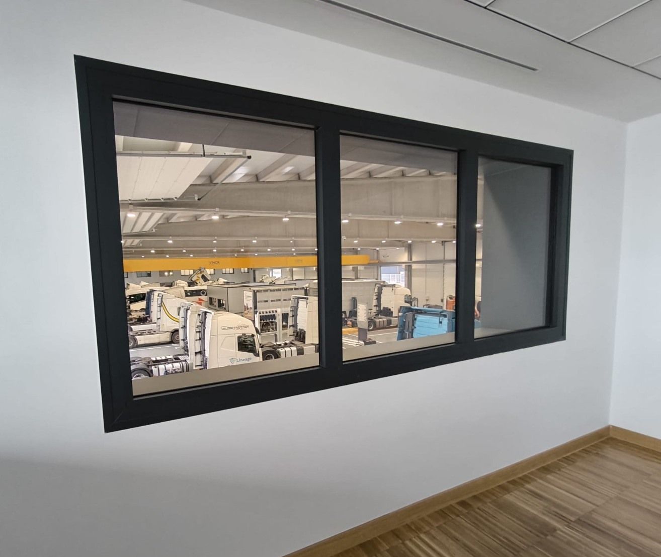 Ventana interior con marco negro que da a un luminoso espacio de trabajo industrial desde un pasillo blanco.