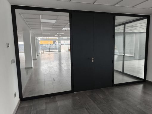 Interior de oficina vacío con paredes de vidrio, puerta oscura y suelo de baldosas que conduce a un espacio abierto.