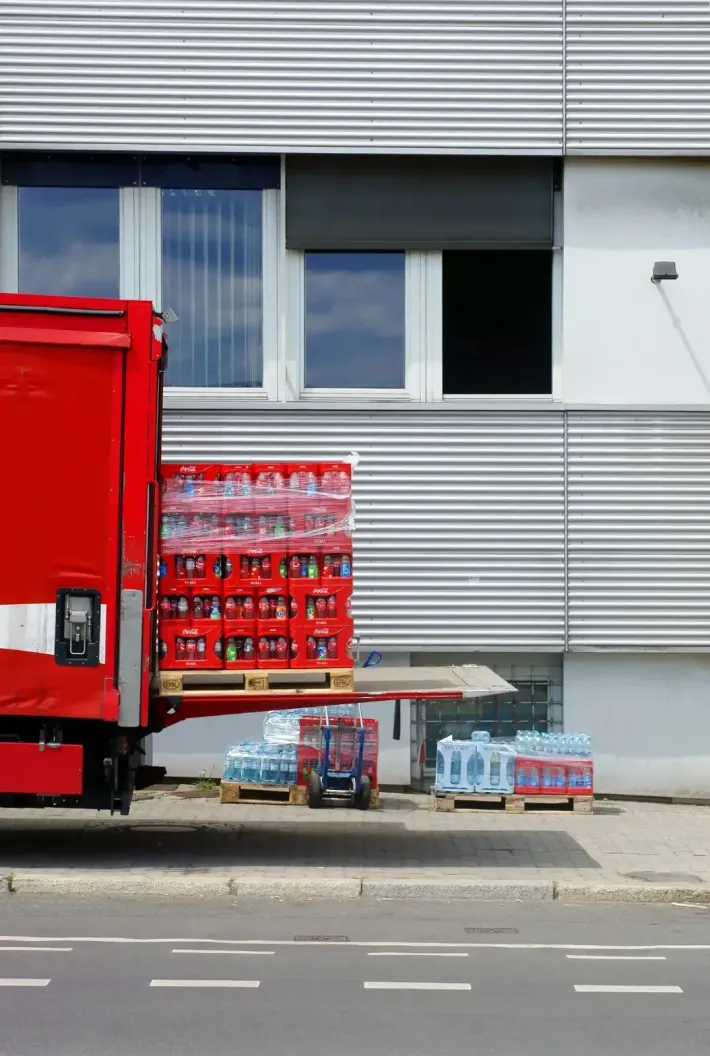 Un camión de reparto rojo estacionado junto a un edificio metálico, con la plataforma elevadora bajada.
