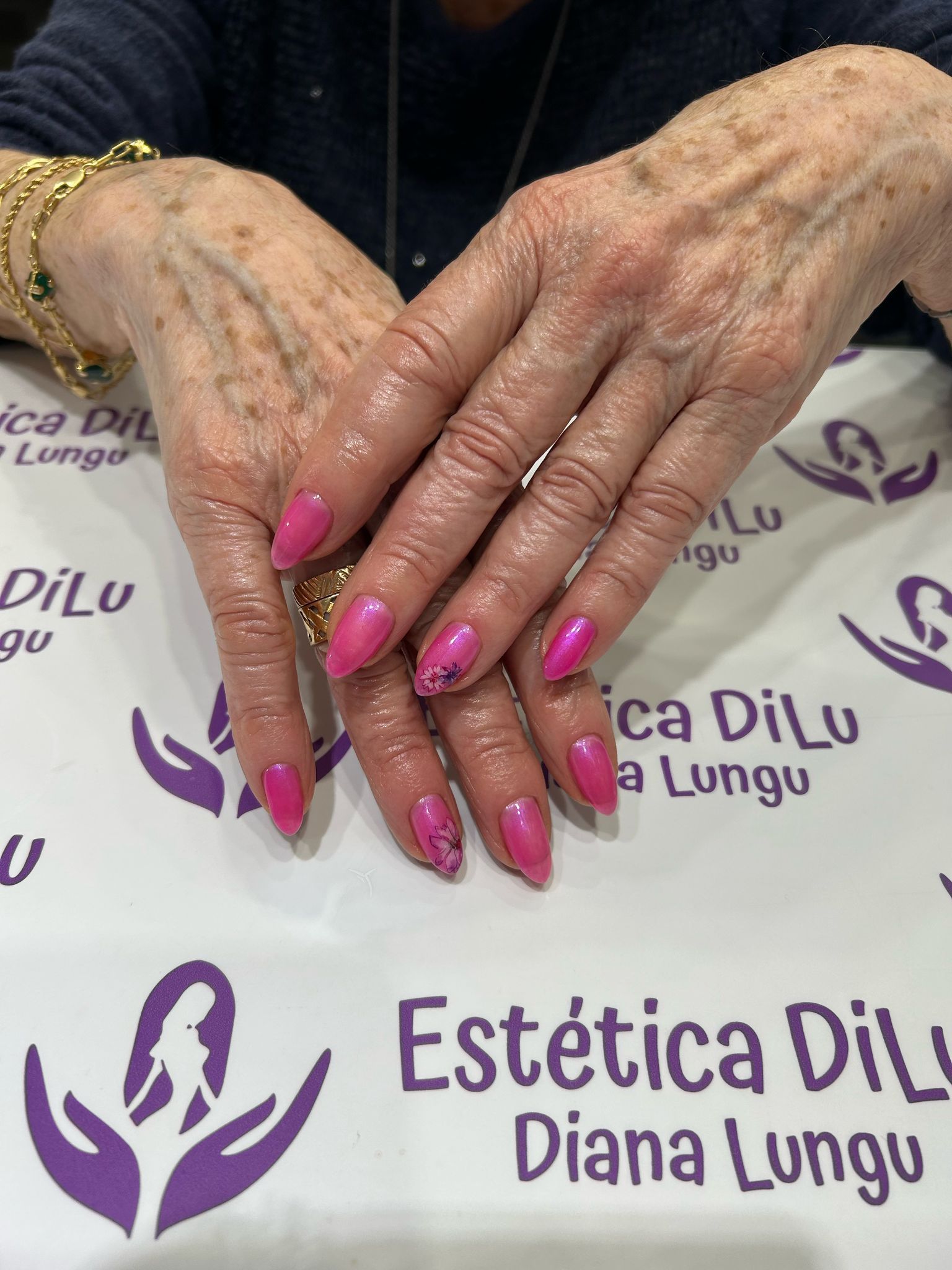 Las manos de una mujer con uñas rosas están sobre una mesa frente a un cartel que dice Estetica Dil Diana Lungu.
