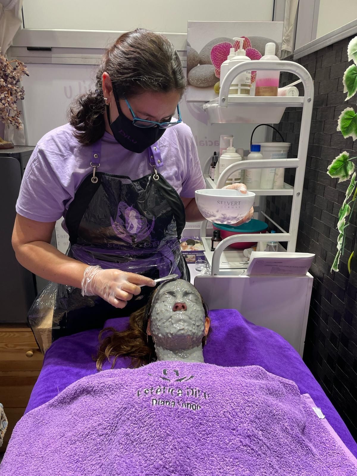 Una mujer que lleva una máscara está aplicando una mascarilla de arcilla en el rostro de una mujer.