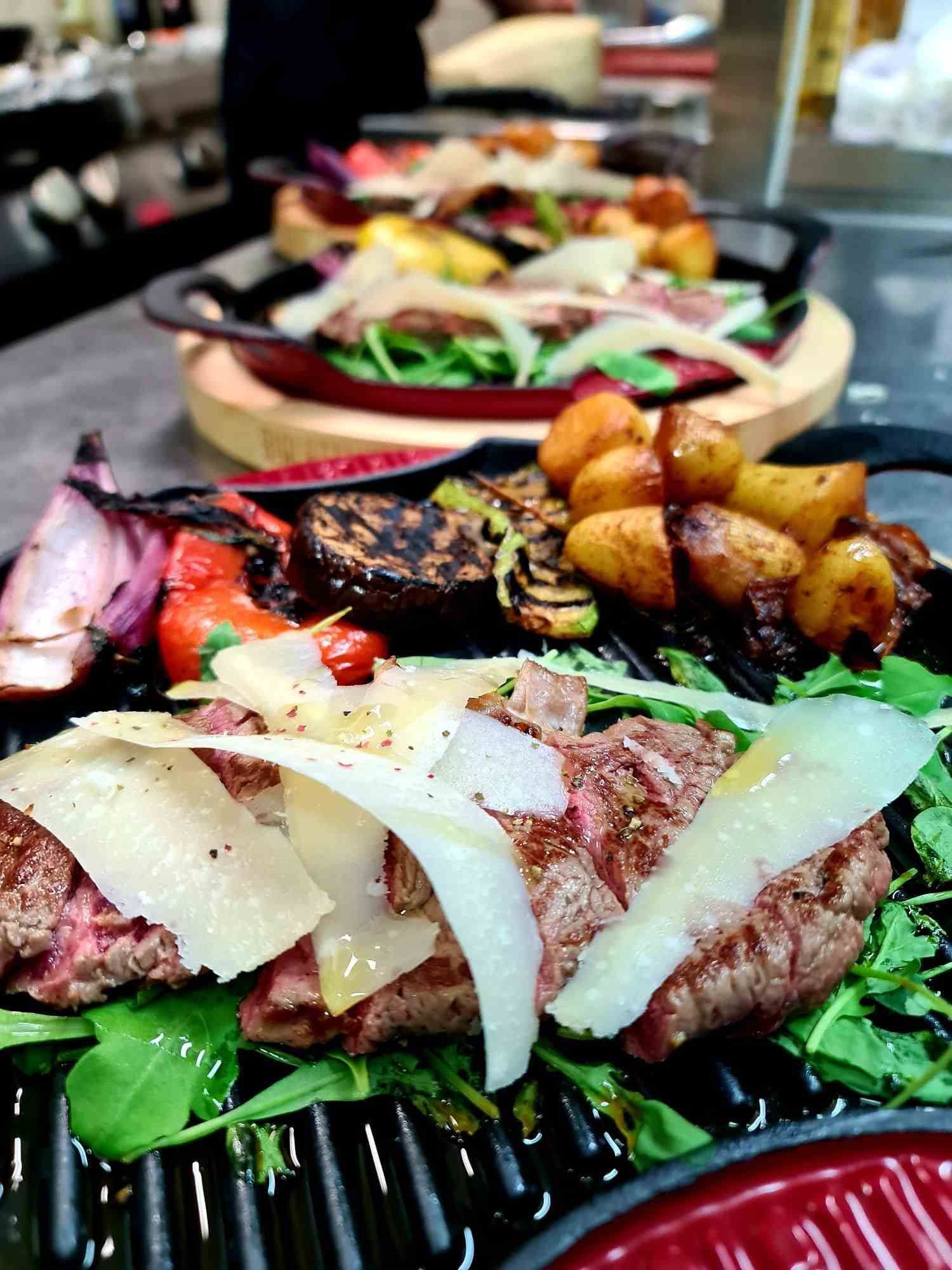 Steak grillé servi avec pommes de terre rôties, légumes, roquette et fromage, sur une plaque chauffante foncée.