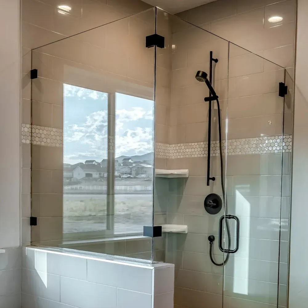 Mampara de ducha de cristal con grifería negra y ventana con vistas a un paisaje.