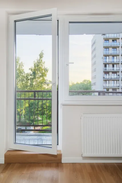 Puerta y ventana blancas abiertas con vista a árboles y un edificio.
