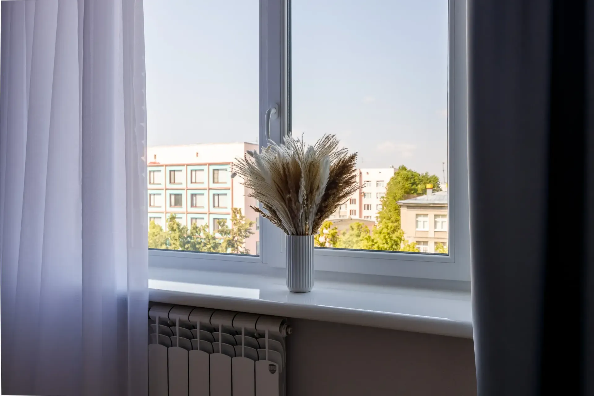 Un jarrón blanco con flores secas en el alféizar de una ventana, con vista a un edificio y árboles.