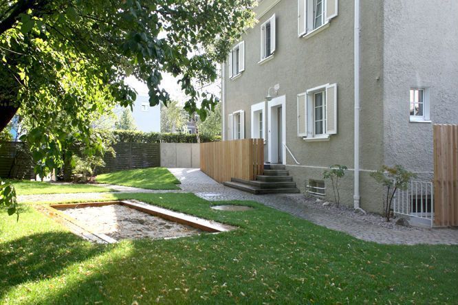 Ein hellgrünes Gebäude mit offenen Fensterläden, ein Holzzaun, eine Rasenfläche mit einem Sandkasten.