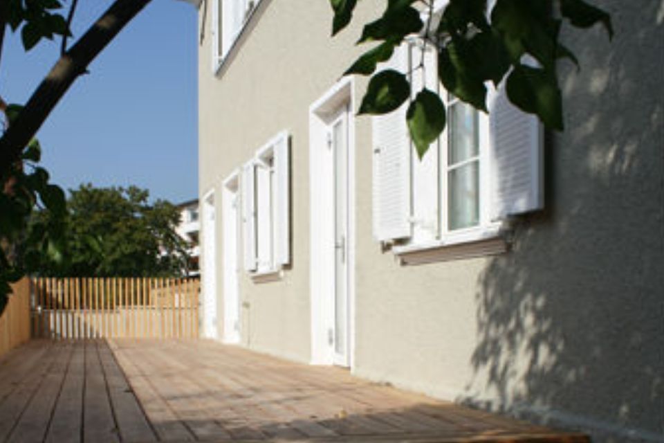 Beigefarbene Gebäudefassade mit weißen Fensterläden, Holzterrasse und einem Ast davor.