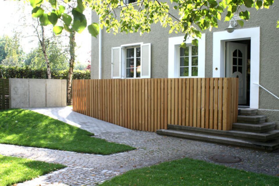 Ein hellbrauner Holzzaun vor einem Haus mit grünem Rasen und Weg.