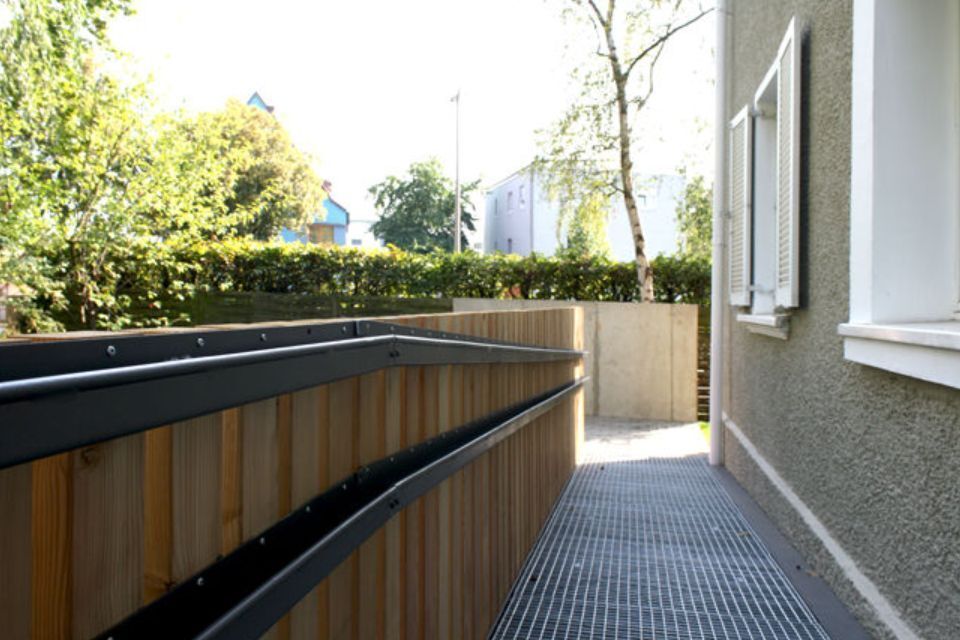Holzrampe mit Metallgeländer neben der grauen Wand und den Fenstern eines Gebäudes, die zu einem Betonbereich führt.