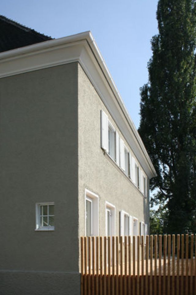 Zweistöckiges Gebäude mit grauen Stuckwänden, weißen Zierleisten, Fenstern und einem Holzzaun; im Hintergrund ein hoher Baum.