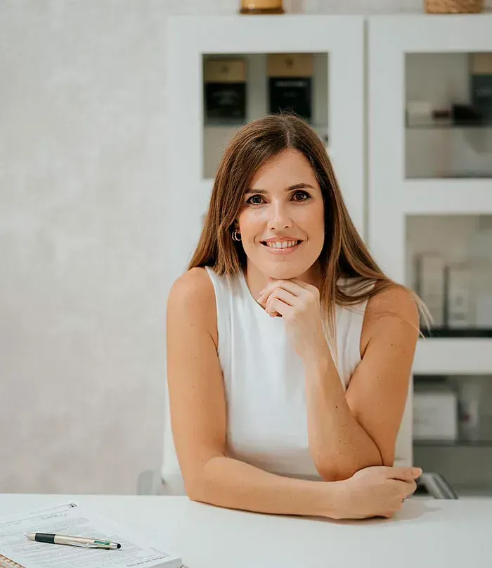 Una mujer de cabello castaño sonríe, apoyando la barbilla en la mano, en un escritorio. Blusa blanca sin mangas, fondo blanco.