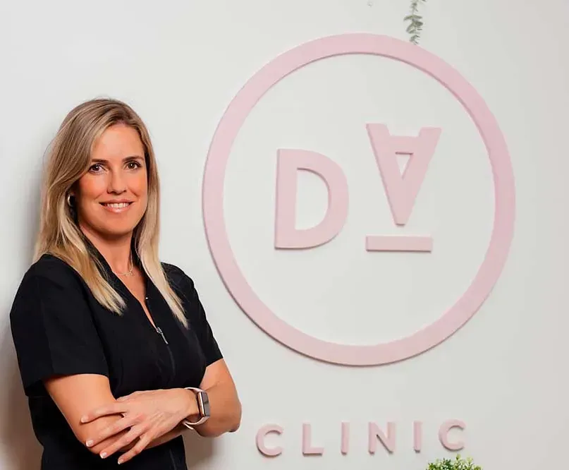 Una mujer con uniforme negro sonríe, con los brazos cruzados, junto a un cartel de clínica rosa con las letras