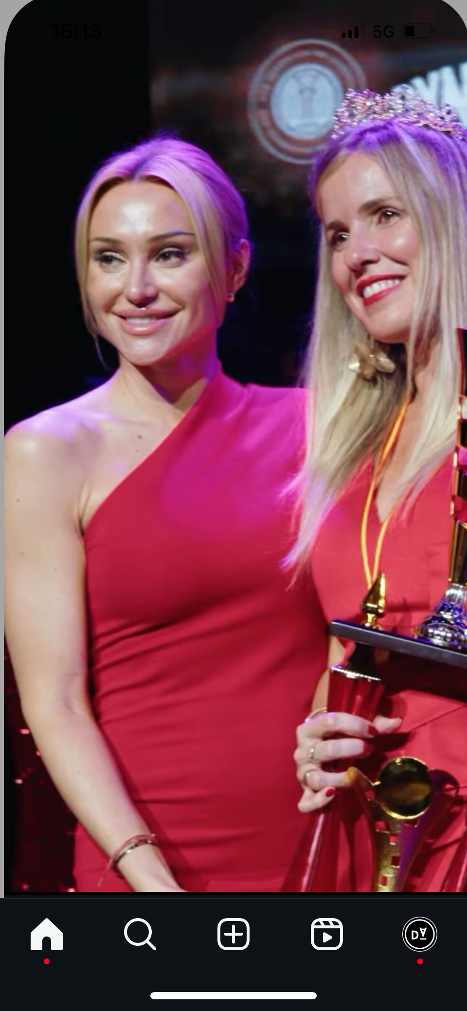 Dos mujeres con vestidos rojos sonriendo, una de ellas sosteniendo un trofeo, en el interior.