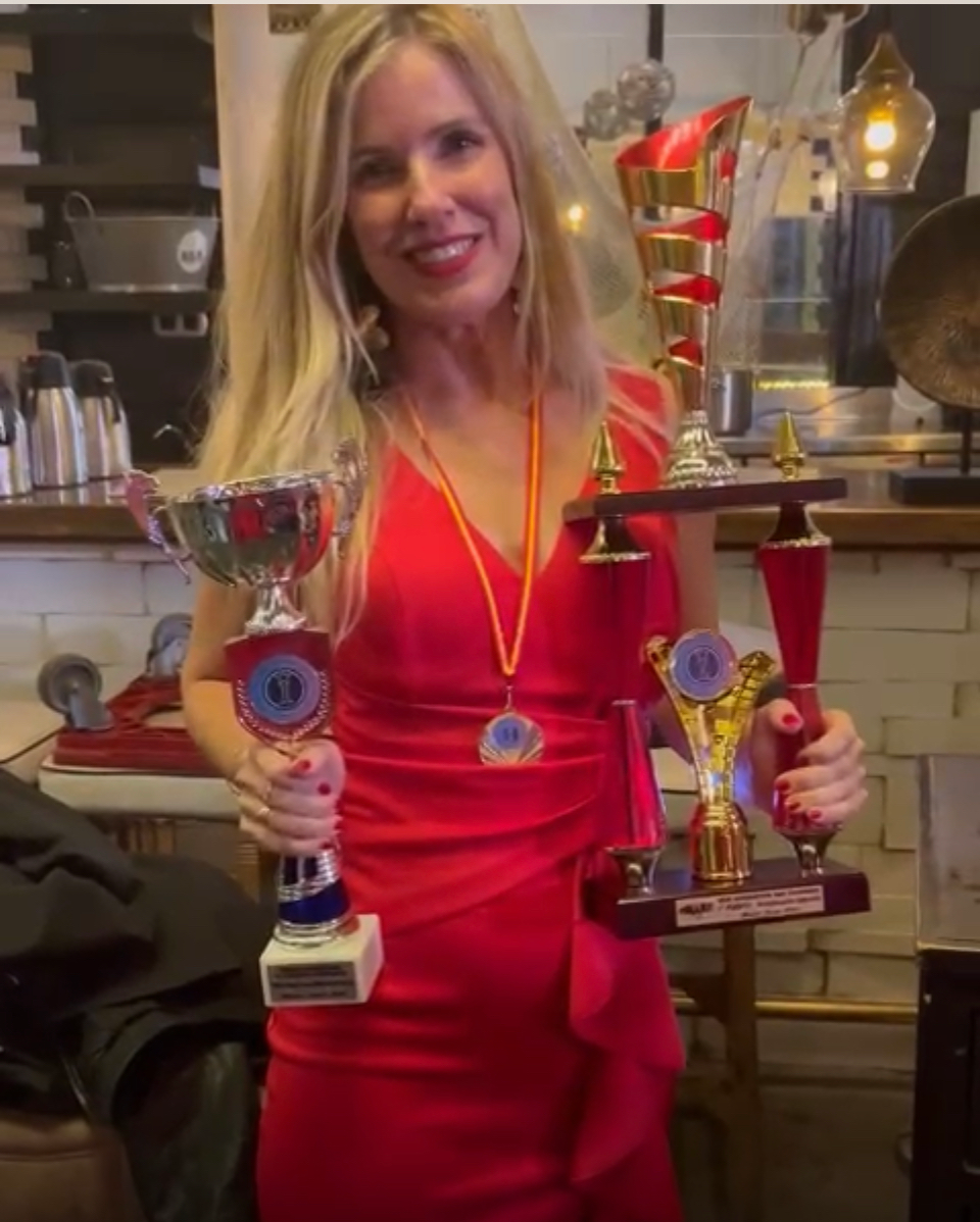 Una mujer vestida de rojo sostiene trofeos, sonriendo en lo que parece ser un restaurante.