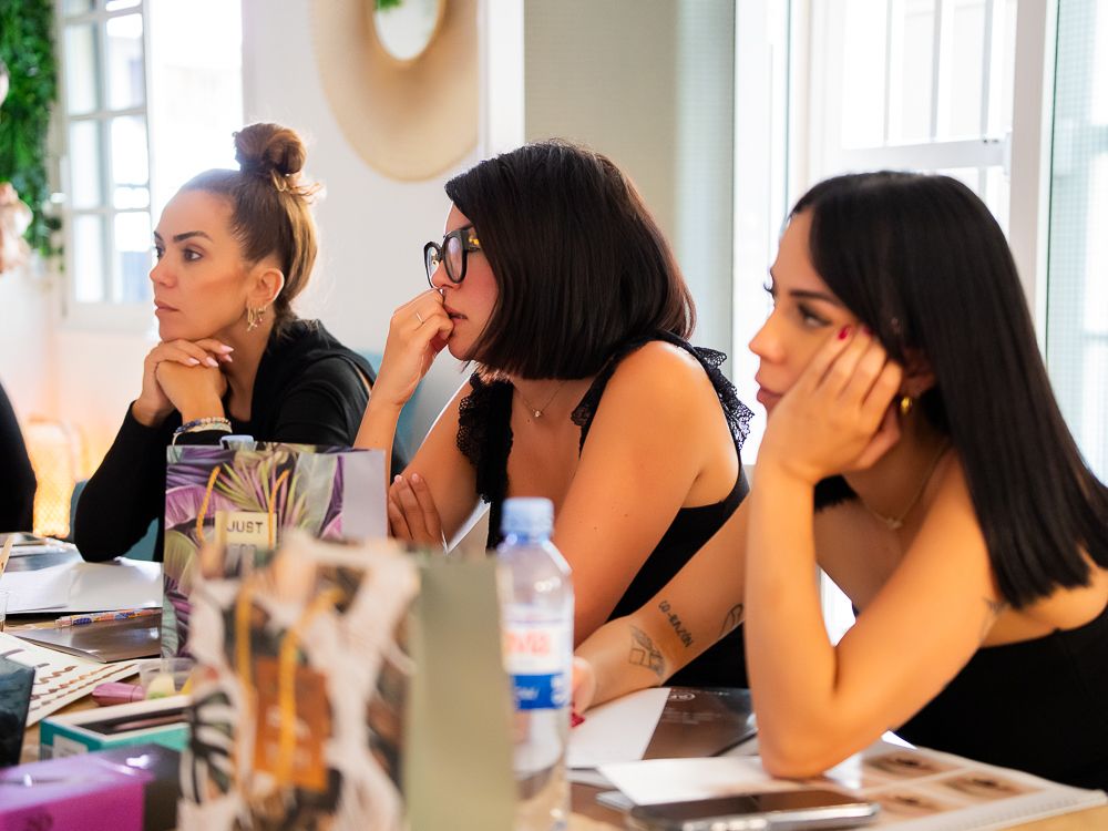 Tres mujeres escuchan atentamente en una mesa, con las manos en la barbilla. Interior luminoso con documentos y una botella de agua.