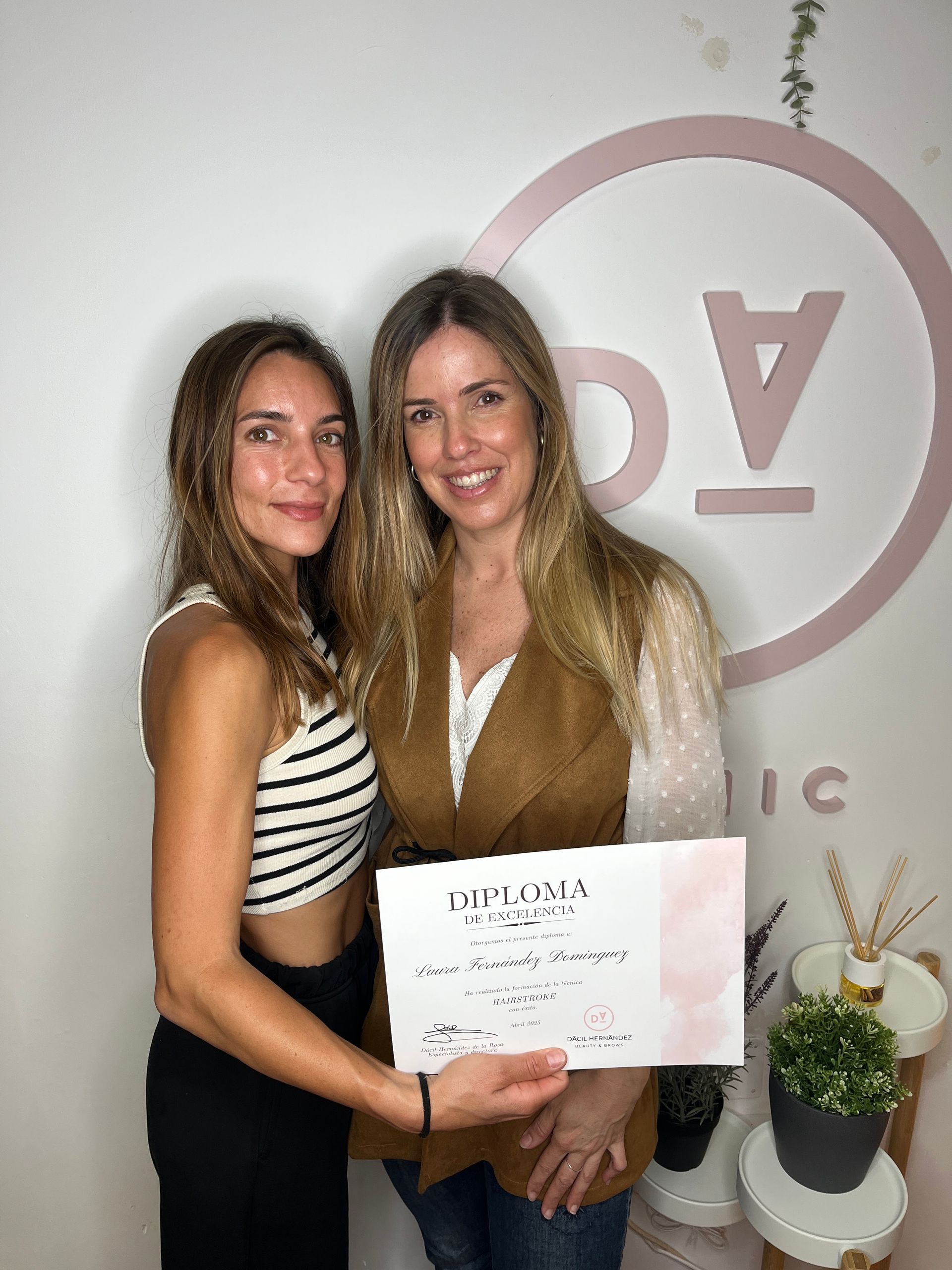 Dos mujeres con un certificado delante de un logotipo. Una con una camiseta de rayas, sonriendo. La otra con un chaleco, sonriendo.