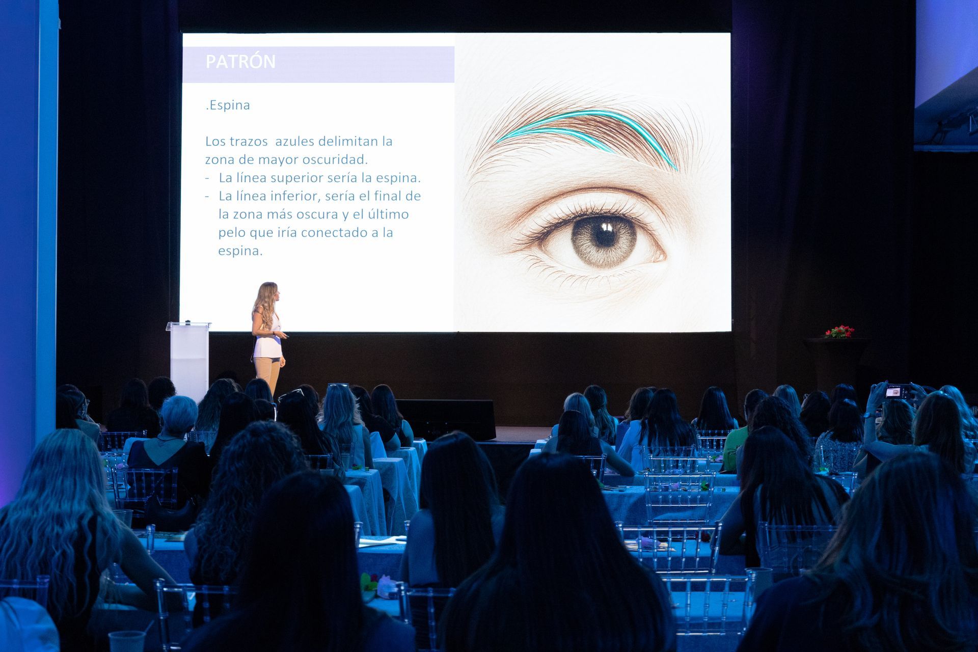 Una mujer se presenta ante el público. Una pantalla detrás de ella muestra un ojo con marcas turquesas.