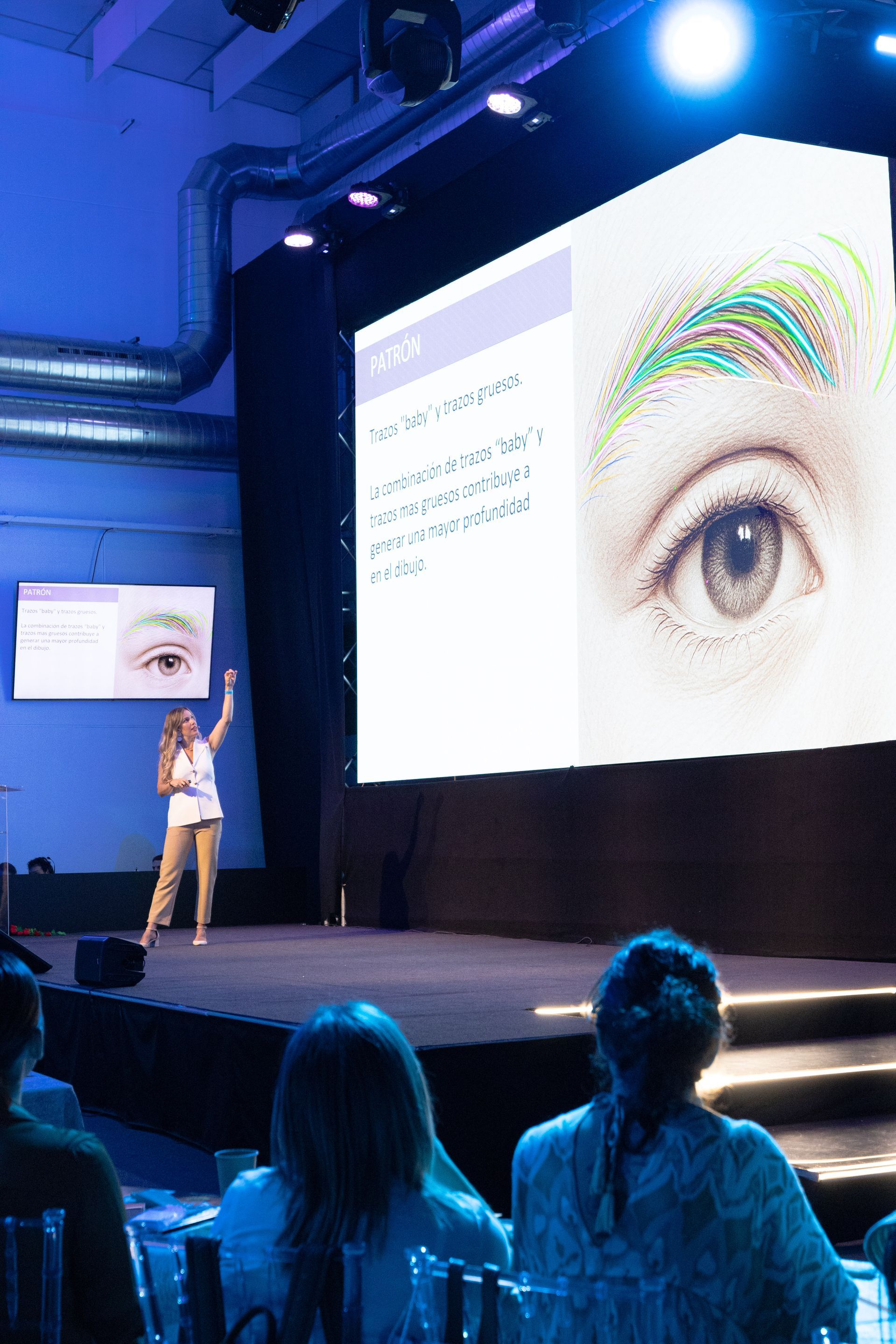 Mujer presentando en una conferencia, señalando una pantalla con un ojo. Público sentado en mesas. Iluminación azul.