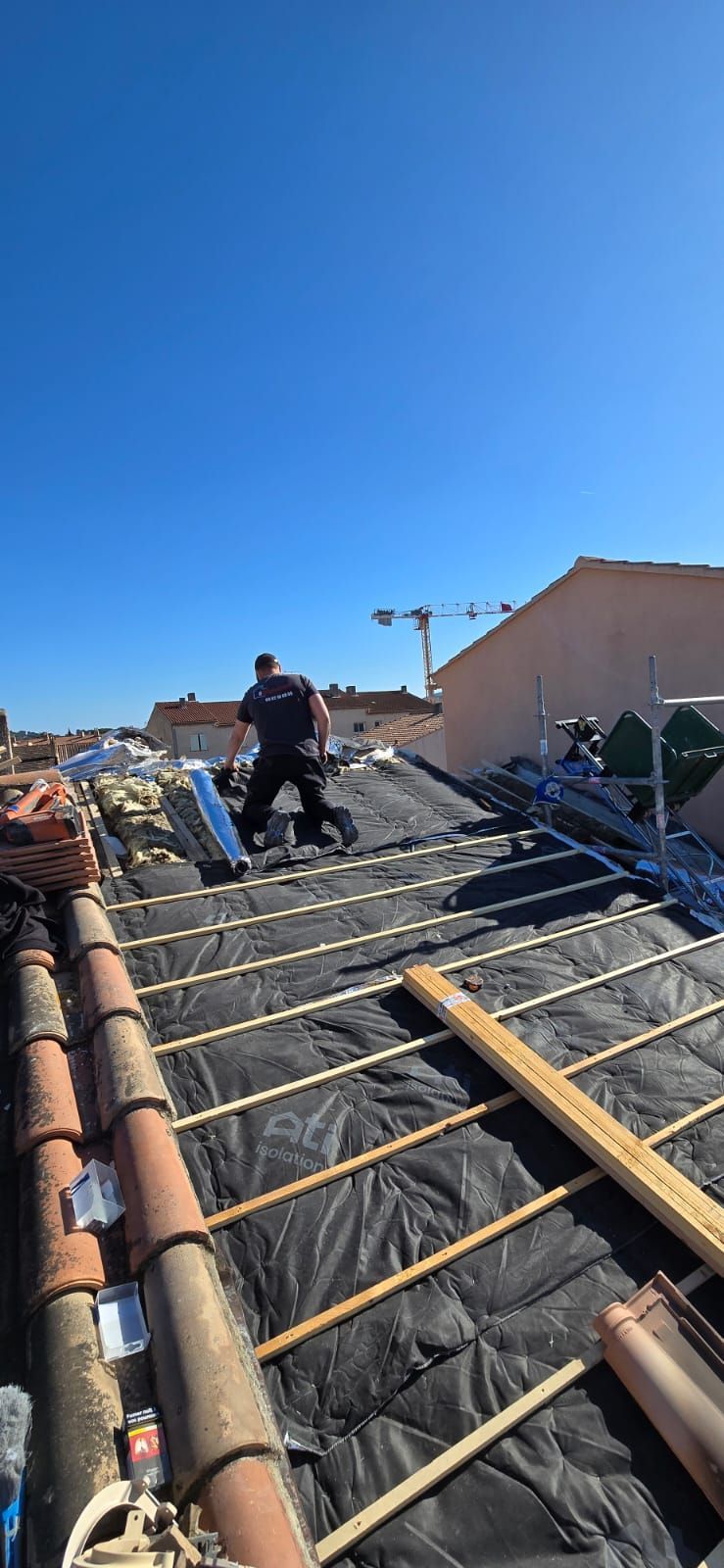 Couvreur travaillant sur un toit en tuiles en terre cuite, avec sous-couche noire, contre un ciel bleu clair.