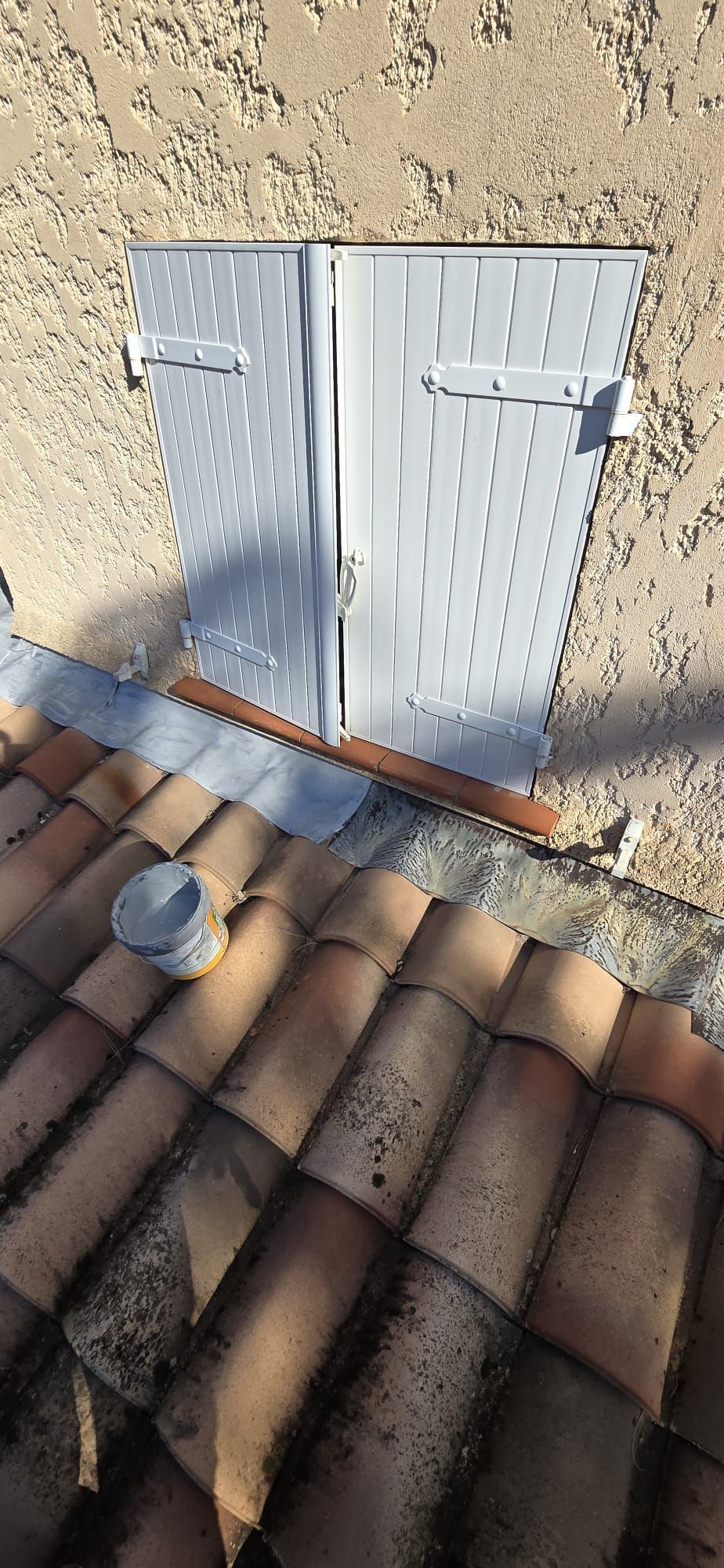 Volets métalliques sur un mur beige au-dessus d'un toit de tuiles rouges, avec un petit seau et une ombre.