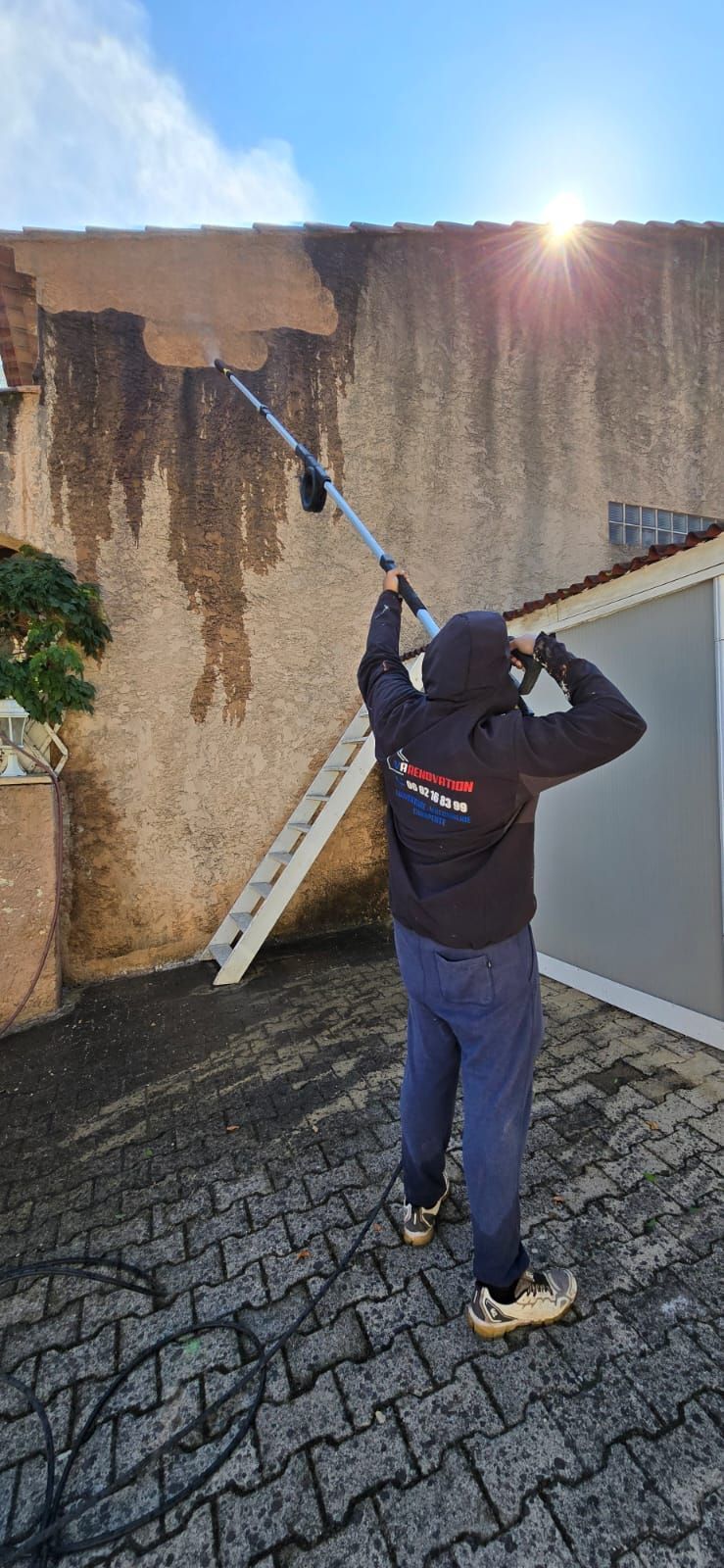 Une personne nettoie un mur à haute pression avec un long outil par une journée ensoleillée.