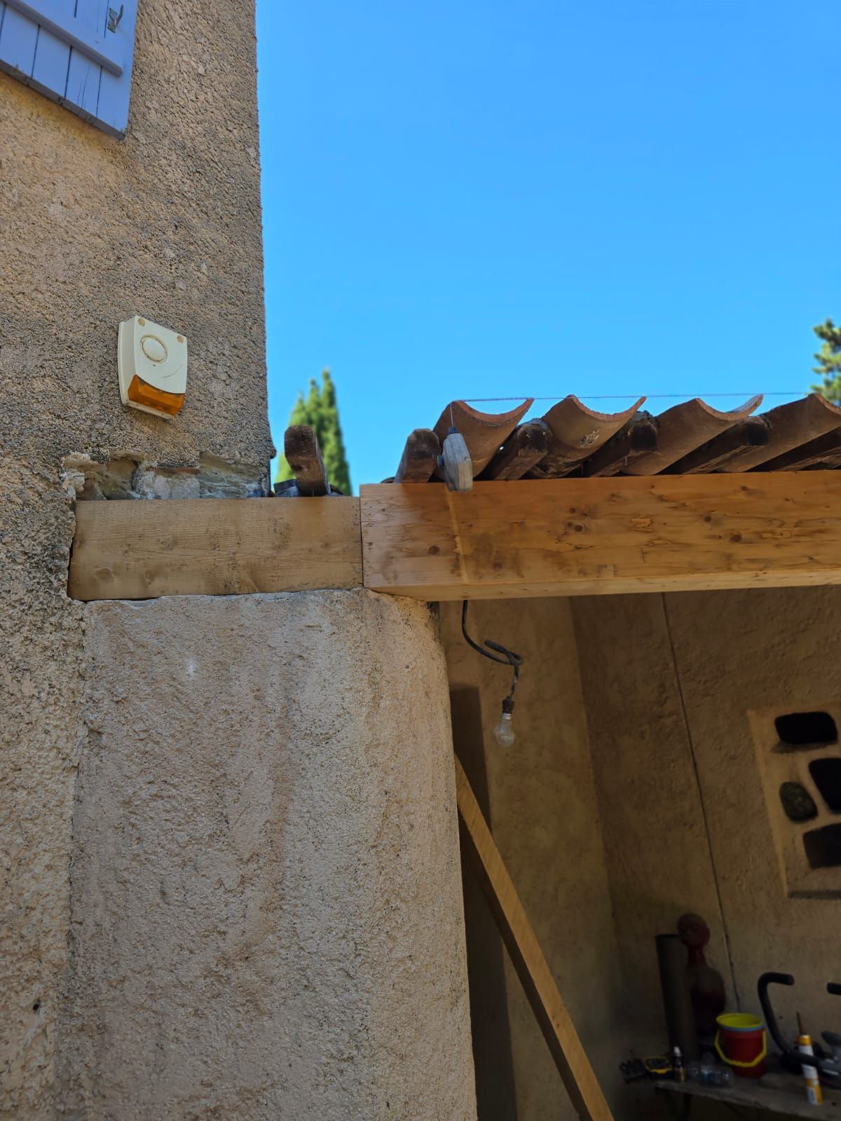 Une poutre en bois patinée soutient un toit en tuiles de terre cuite, fixé à un bâtiment en stuc sous un ciel bleu.