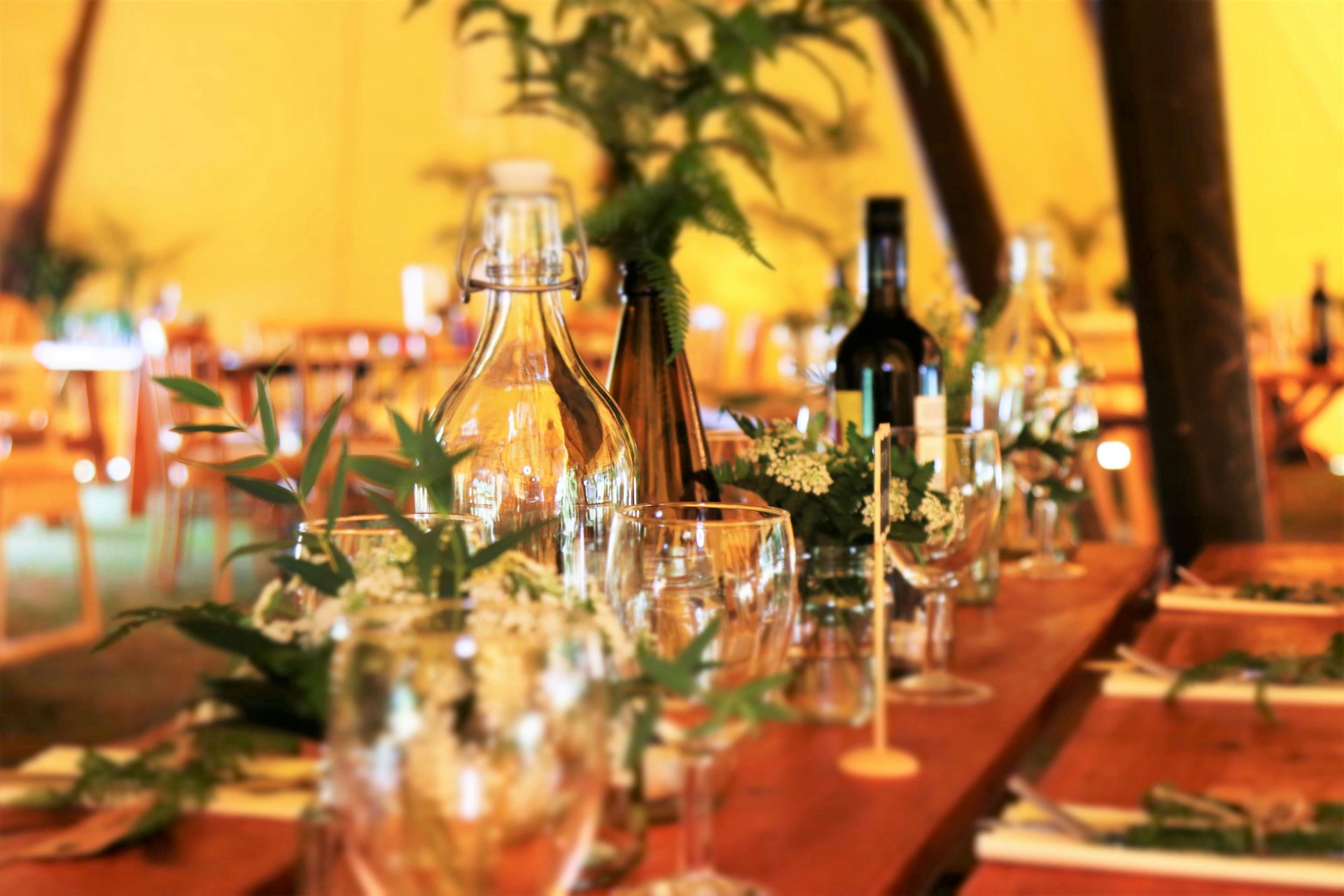Table setting with bottles, glasses, and greenery in a tent. Wooden table and warm lighting.