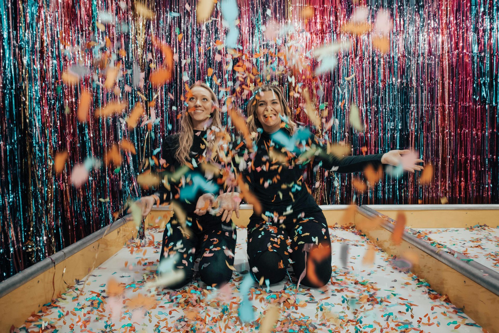 Two women kneeling in confetti, smiling, in front of a shiny curtain backdrop.