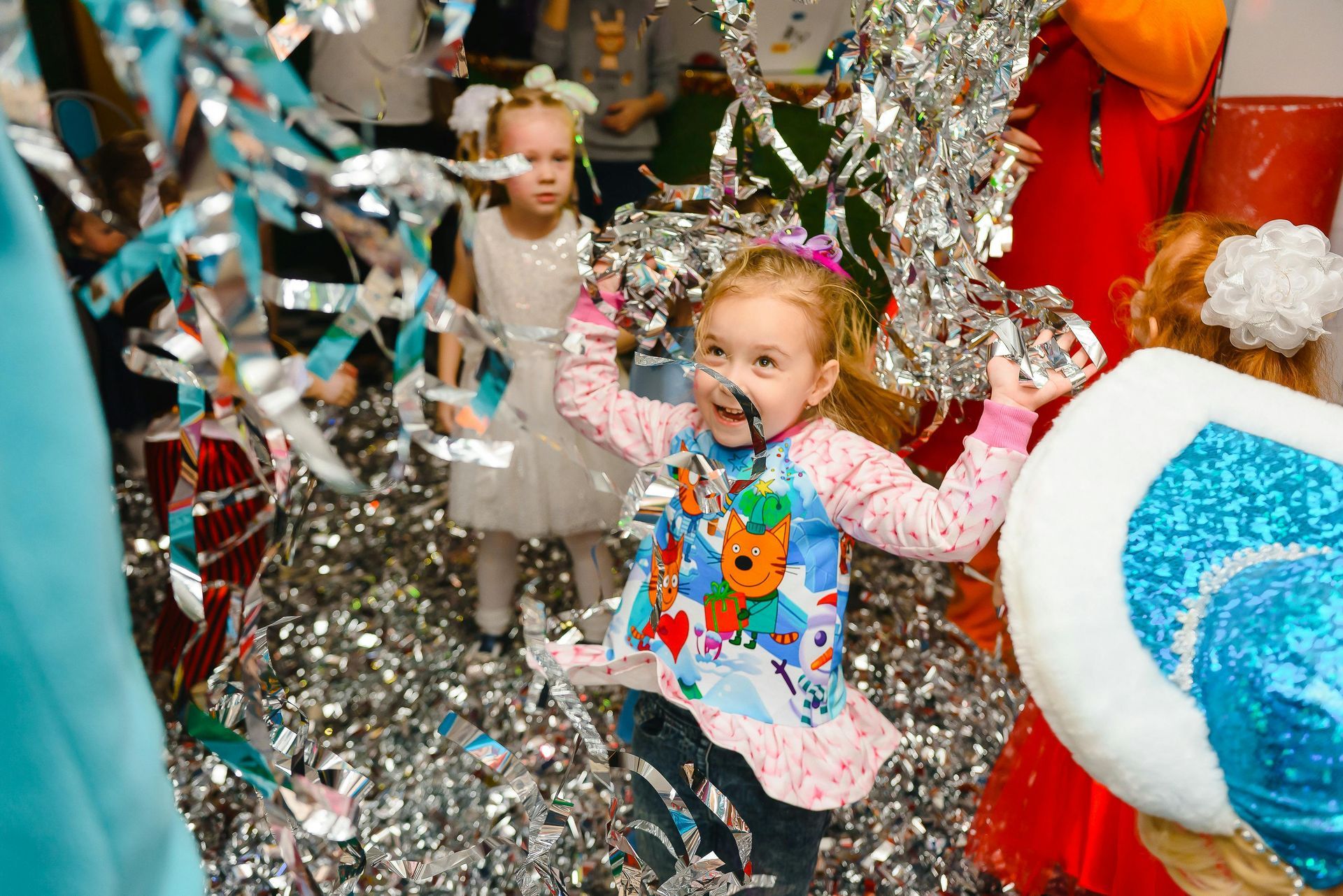 Girl in a festive setting throwing silver confetti with joyful expression, surrounded by other children.