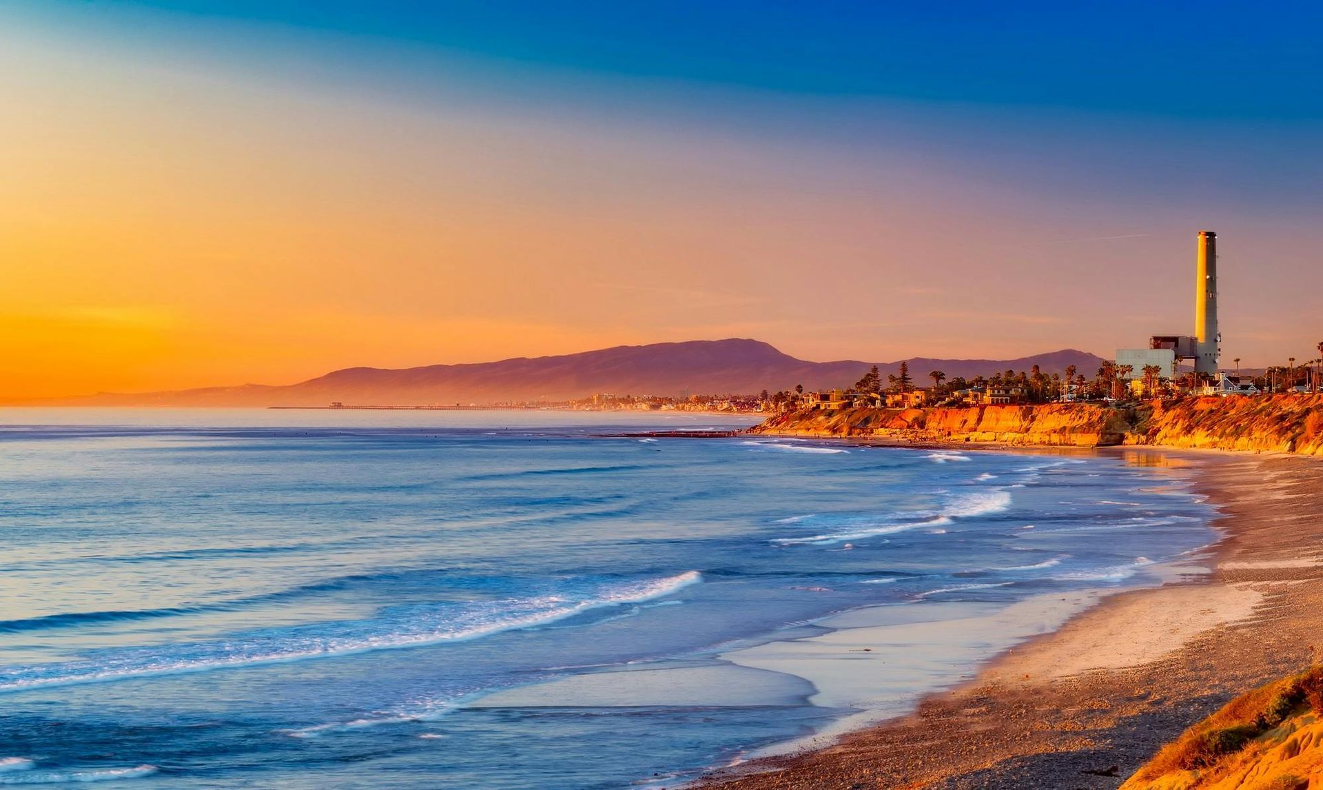 Ocean waves lapping on a beach at sunset, with a tall industrial smokestack visible.