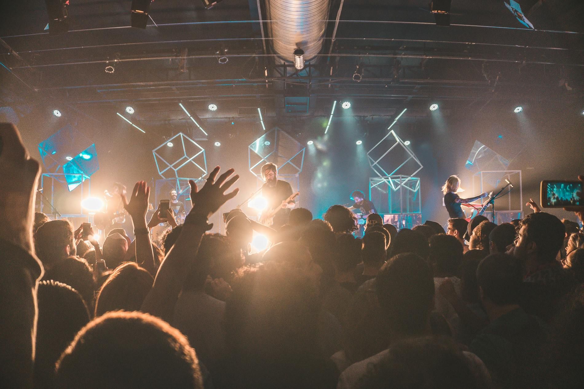 Concert crowd with raised arms, backlit by stage lights. Musicians on stage, performing.