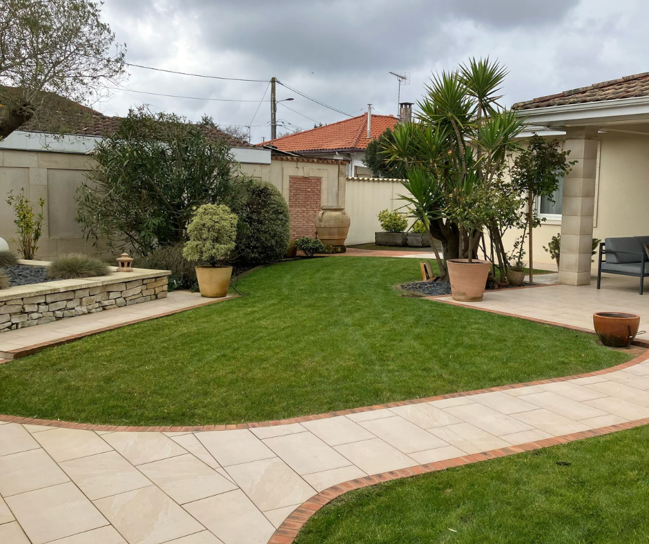 Cour arrière bien entretenue avec une terrasse en pierre, une pelouse verte et diverses plantes en pot sous un ciel nuageux.