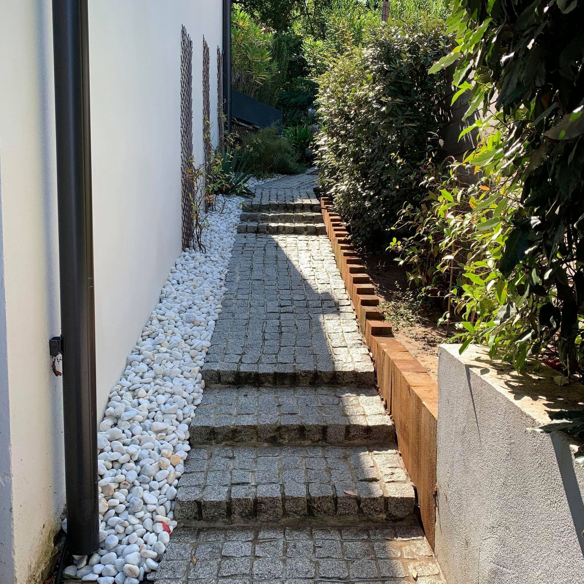 Des marches en pierre montent entre un mur blanc orné de galets blancs et de verdure, avec un mur de soutènement sur la droite.