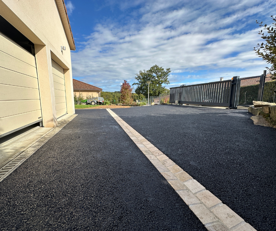 Allée en asphalte noir bordée de pierres beiges, menant à un garage et un portail automatique, sous un ciel bleu.