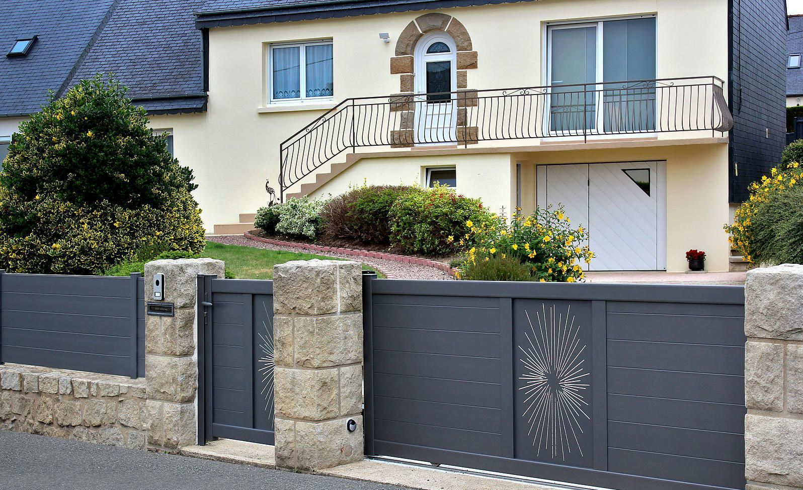 Clôture et portail modernes gris devant une maison jaune clair avec balcon et escalier, arbustes.