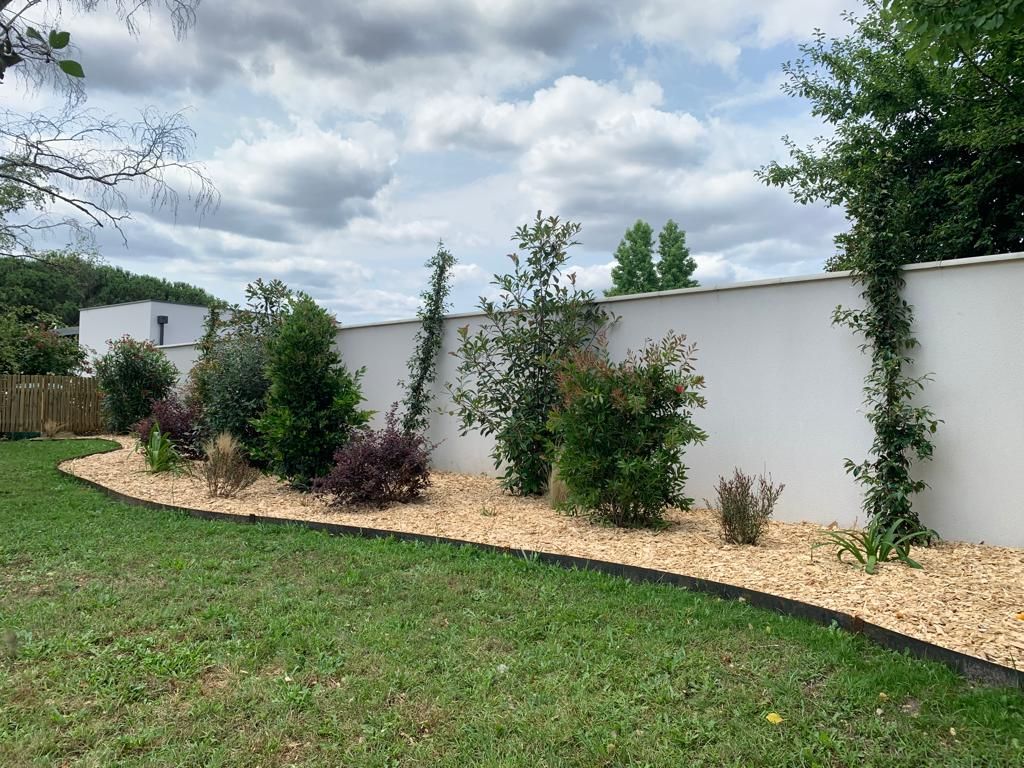 Un jardin paysager avec un mur blanc et une végétation variée, bordé de gravier brun et de bordures noires.