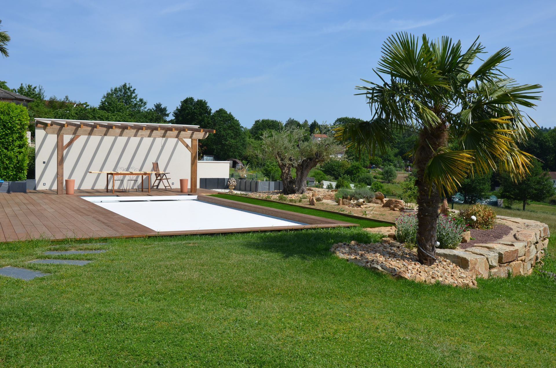 Jardin avec piscine, pergola, terrasse en bois et massif de pierre avec palmier et autres végétaux.