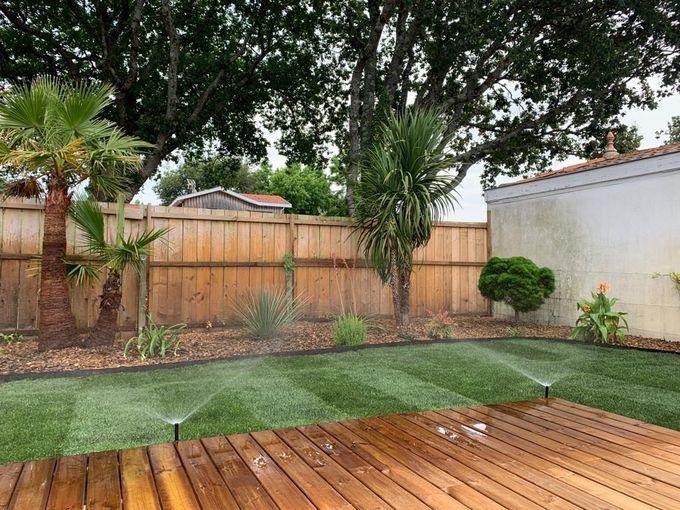 Scène de jardin avec système d'arrosage automatique arrosant la pelouse ; terrasse en bois au premier plan, clôture en bois et arbres à l'arrière-plan.