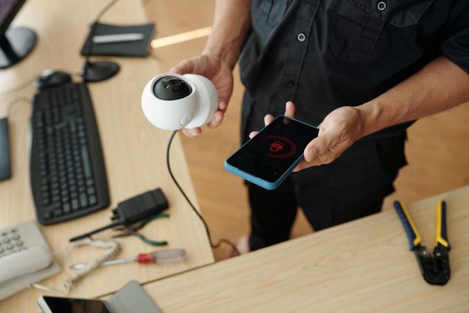 Persona sosteniendo un teléfono inteligente con un ícono de alerta roja y una cámara de seguridad blanca sobre un escritorio 