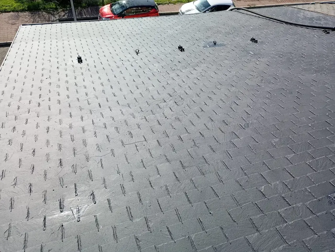 Tapete negro texturizado con numerosos agujeros pequeños dispuesto sobre una superficie, con coches al fondo.