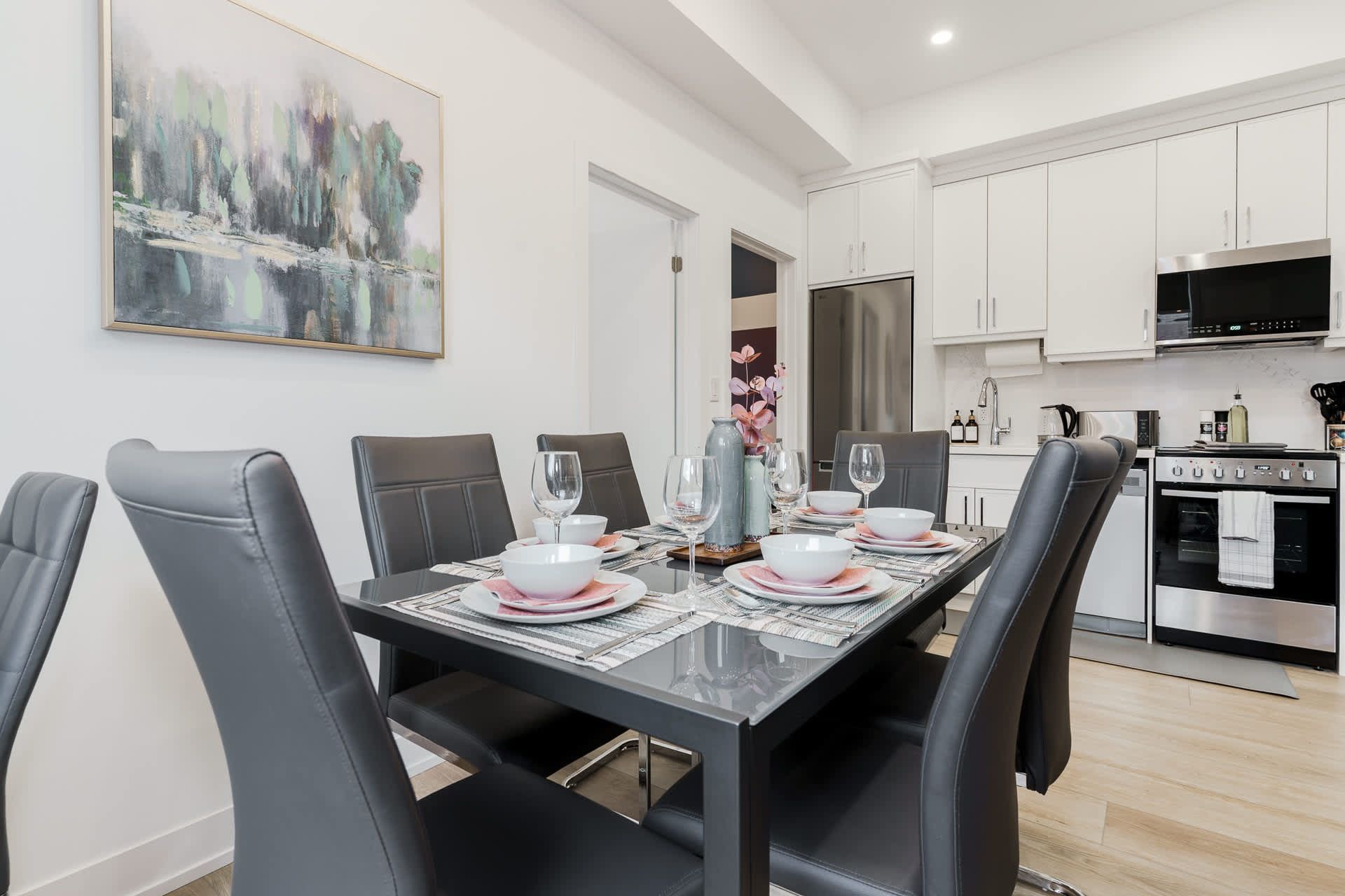 Dining room with a table set for a meal; a painting hangs on the wall, and kitchen appliances are visible.