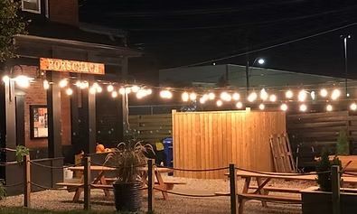 Outdoor patio at night with string lights, picnic tables, and wooden fence. The sign says 
