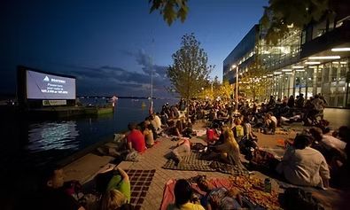 Outdoor movie screening by a lake; people seated on blankets, watching a screen; night.
