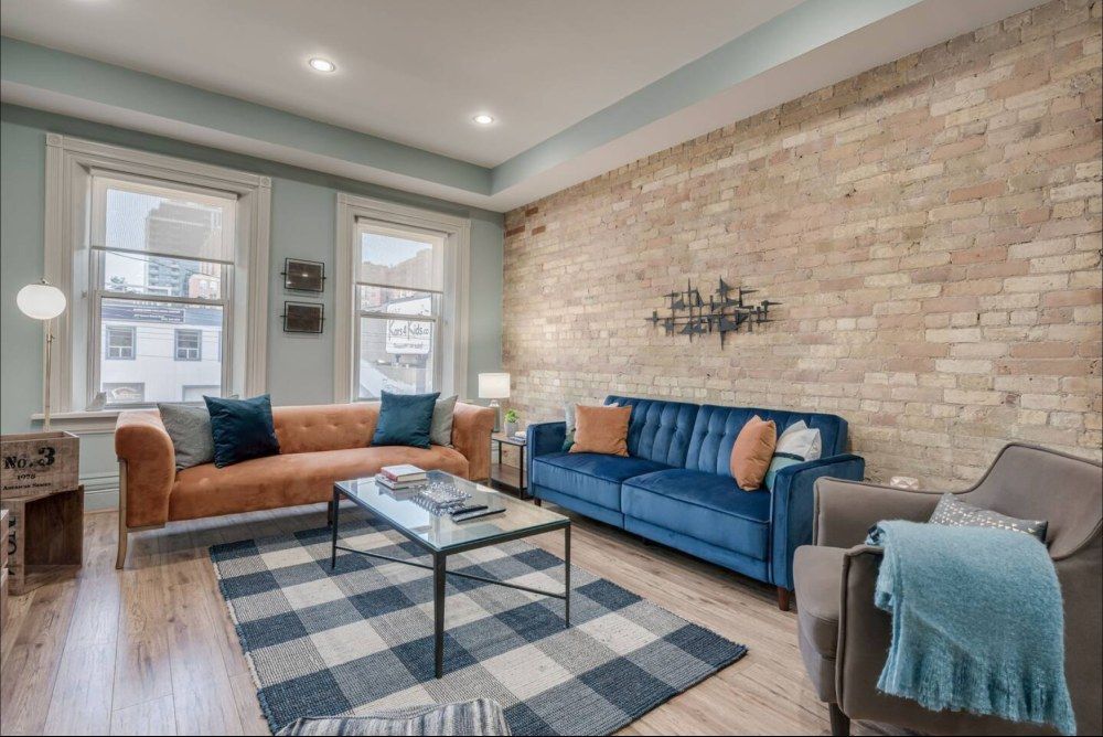 Living room with orange and blue sofas, exposed brick wall, checkered rug, windows.