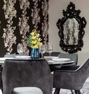 Dining room with floral wallpaper, black chairs, and ornate mirror. Table set with glasses and flowers.