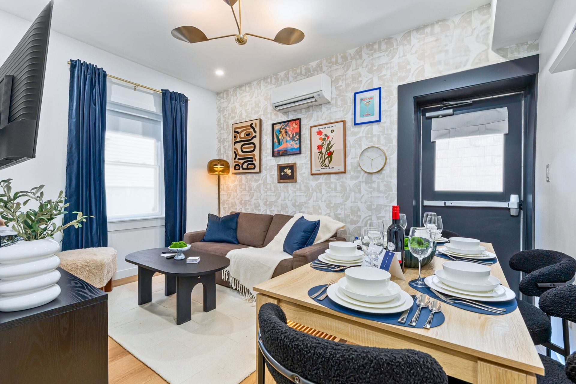 Cozy living room with beige sofa, patterned wallpaper, blue curtains, framed art, and a set dining table