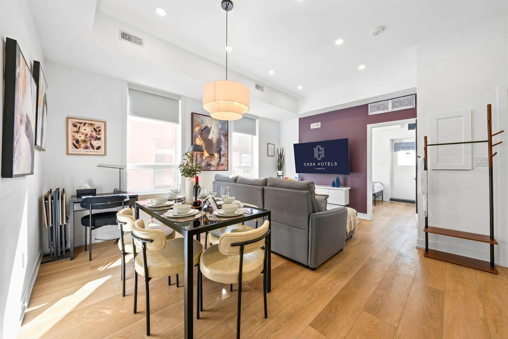 Bright modern dining and living room with gray sofa, wall-mounted TV, wooden floors, and large windows.