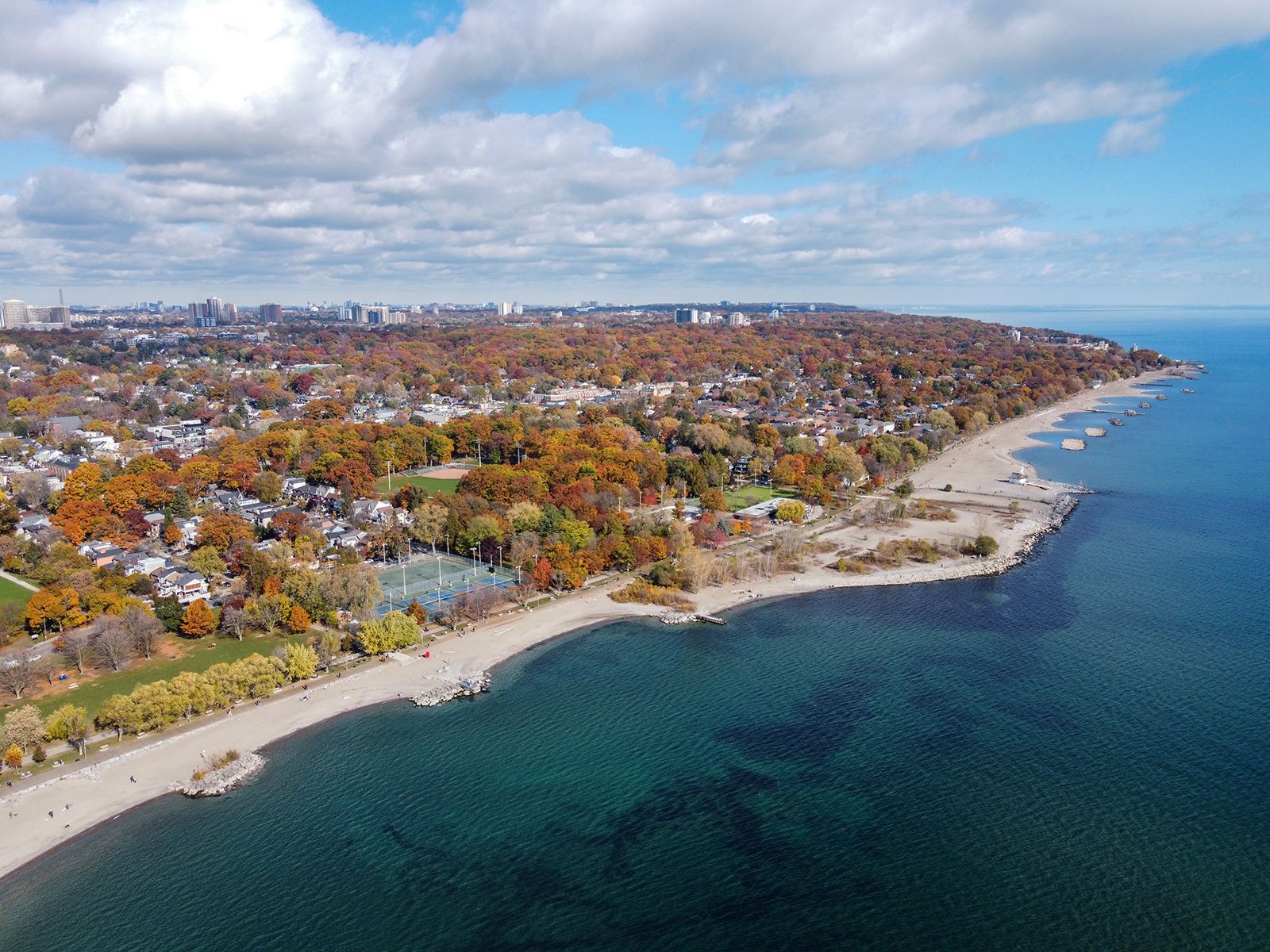 Aerial view of beach, park, and city skyline under a cloudy sky, with fall foliage in shades of orange and brown.